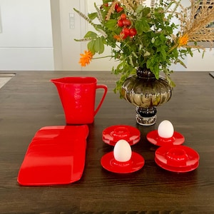 May include: A set of red plastic egg cups and a red plastic tray with a red plastic pitcher in the background. The egg cups are filled with white eggs.