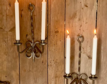 May include: Two pairs of wrought iron candle sconces with lit white candles. The sconces are mounted on a wooden wall. Each sconce holds two candles and has a decorative, swirling design. The candle flames are bright.
