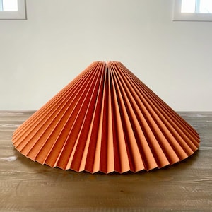 May include: A large, pleated lampshade in a burnt orange colour. The shade is made of a fabric material and has a wide, circular shape.