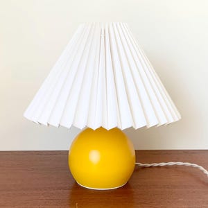 May include: A yellow ceramic table lamp with a white pleated lampshade. The lamp has a cord and plug.