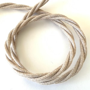 May include: A coiled, beige twisted electrical cord. The cord is made of a woven fabric and has a textured appearance. The neutral color and twisted design suggest a decorative or vintage style.