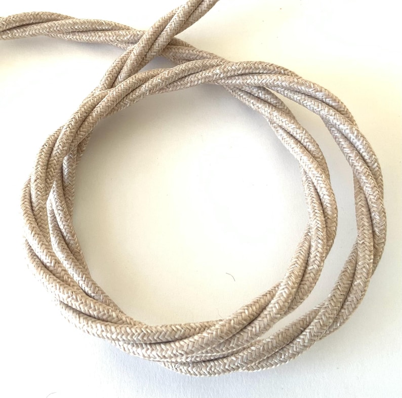 May include: A coiled, beige twisted electrical cord. The cord is made of a woven fabric and has a textured appearance. The neutral color and twisted design suggest a decorative or vintage style.
