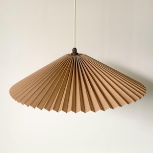 Op de afbeelding: Een bruin, geplooid papieren lampenkap met een conische vorm, opgehangen aan een wit snoer. De lampenkap heeft een bronzen armatuur aan de bovenkant. Het ontwerp heeft verticale plooien.