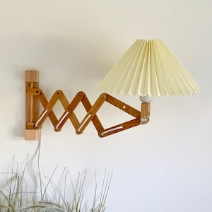 Puede incluir: Una l&aacute;mpara de pared de madera con una pantalla de tela blanca plisada. La l&aacute;mpara tiene un brazo plegable y es ajustable.