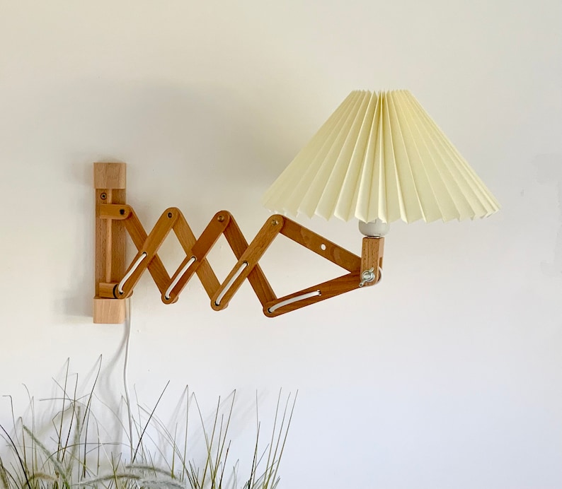 Puede incluir: Una l&aacute;mpara de pared de madera con una pantalla de tela blanca plisada. La l&aacute;mpara tiene un brazo plegable y es ajustable.