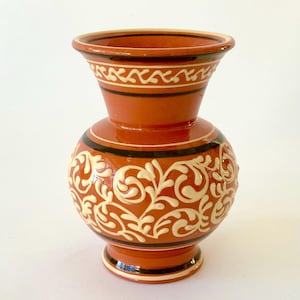 May include: A terracotta-colored ceramic vase with a bulbous body and flared top. The vase features cream-colored floral patterns and black and white bands. The vase is approximately 7 inches tall.