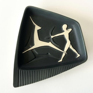 May include: A dark charcoal gray ceramic tray with an irregular trapezoidal shape. The center features a raised, cream-colored relief design of a stylized archer aiming a bow at a leaping deer. The bottom edge has a textured, vertical ribbed pattern.