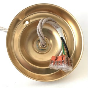 May include: A brass-colored ceiling canopy with a central threaded hole and a beige braided cord. Electrical wires, including black, white, and green, are connected with orange wire connectors. The canopy has a circular shape.