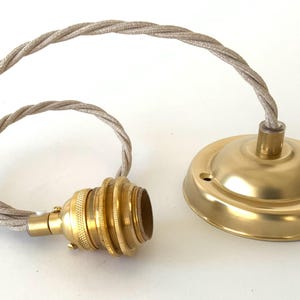 May include: A brass-colored pendant light fixture with a twisted, beige fabric cord. The fixture includes a bulb socket and a ceiling canopy. The components are arranged on a white surface.