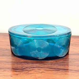 May include: A teal-colored, oval-shaped glass container with a lid. The lid and sides feature a concentric circle design. The container is sitting on a wooden surface.