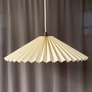 Könnte beinhalten: Cremefarbene, plissierte Papier-Pendelleuchte. Der Schirm hat eine breite, konische Form mit einem gewellten Rand. Die Leuchte hängt an einem weißen Kabel und hat eine bronzefarbene Metallkappe.