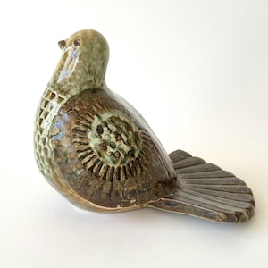 Könnte beinhalten: Eine Keramik-Vogelfigur mit einer glänzenden Glasur in Olivgrün- und Dunkelbrauntönen. Der Körper zeigt ein strukturiertes, wabenartiges Muster auf der Brust und ein kreisförmiges, sonnenstrahlenartiges Design auf dem Flügel sowie einen flachen Schwanz.
