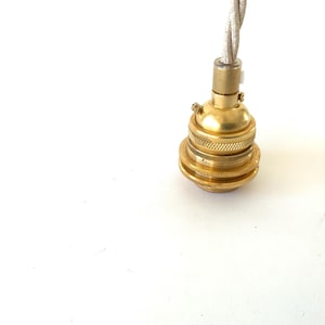 May include: A close-up of a brass-colored light socket with a decorative, ridged design. A twisted, off-white fabric-covered wire extends from the top. The socket is designed for a light fixture.
