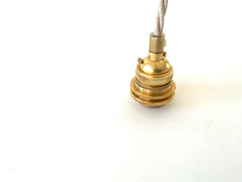 May include: A close-up of a brass-colored light socket with a decorative, ridged design. A twisted, off-white fabric-covered wire extends from the top. The socket is designed for a light fixture.