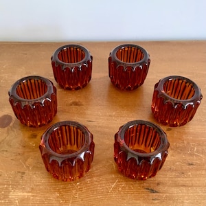May include: A set of six amber-coloured glass votive candle holders, each featuring a cylindrical shape with vertical ribbed detailing along the exterior walls and a smooth, circular opening at the top for holding small candles or tea lights.
