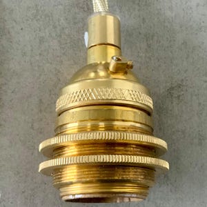 May include: Close-up of a brass-colored light socket with a textured design. The socket has a threaded base and a gold-colored braided cord. The light fixture is designed for use in a lamp or other lighting project.