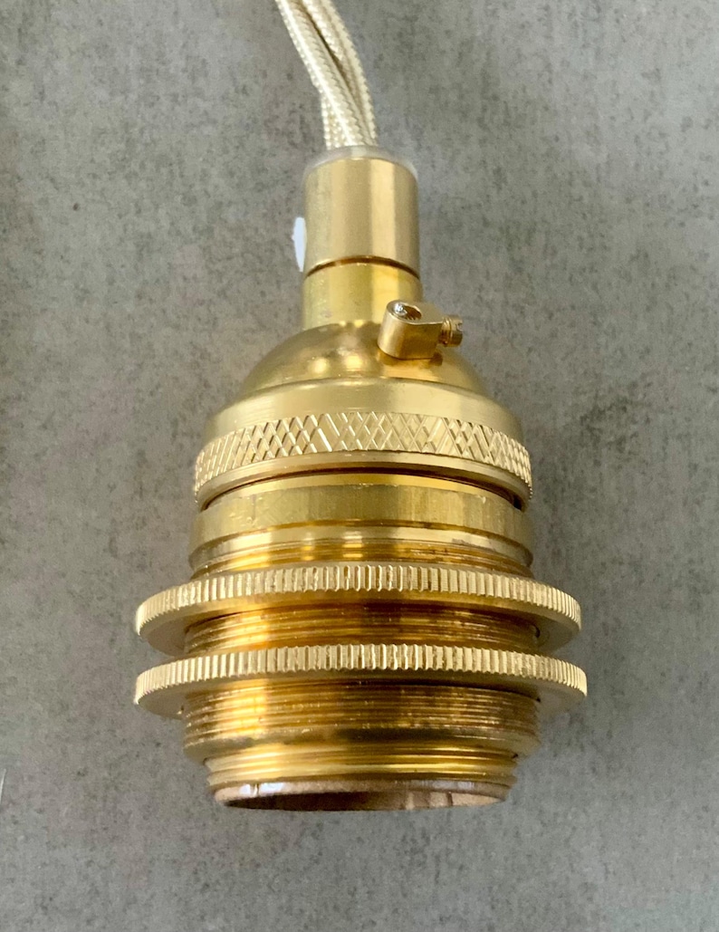 May include: Close-up of a brass-colored light socket with a textured design. The socket has a threaded base and a gold-colored braided cord. The light fixture is designed for use in a lamp or other lighting project.