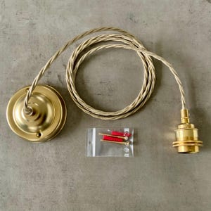 May include: A brass-colored pendant light fixture kit. The kit includes a ceiling canopy, a light socket, a twisted fabric-covered cord, and a small bag of mounting hardware. The components are arranged on a gray surface.