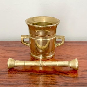 Könnte beinhalten: Ein Vintage-Mörser und Stößel aus Messing. Der Mörser hat einen ausgestellten Rand, zwei Griffe und einen abgerundeten Boden. Der Stößel ist zylindrisch mit abgerundeten Enden. Das Set wird auf einer Holzoberfläche präsentiert.