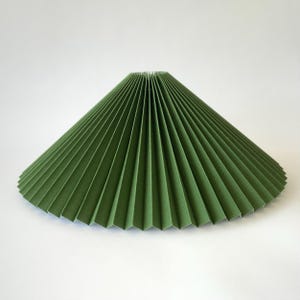 May include: A green pleated lampshade with a conical shape. The shade is made of paper or fabric and has a series of vertical folds that create a textured surface. The shade is designed to diffuse light and add a decorative element to a lamp.