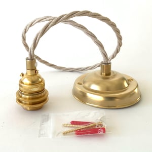 May include: A brass-colored pendant light fixture with a matching ceiling canopy and a twisted beige fabric cord. Includes a clear plastic bag with red and gold screws and wall anchors. The fixture is designed for easy installation.