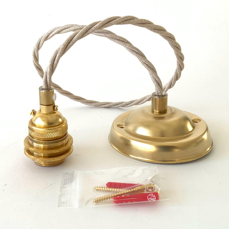 May include: A brass-colored pendant light fixture with a matching ceiling canopy and a twisted beige fabric cord. Includes a clear plastic bag with red and gold screws and wall anchors. The fixture is designed for easy installation.