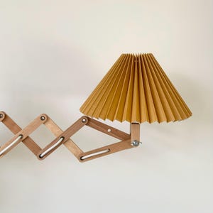 Puede incluir: Una l&aacute;mpara de pared de madera con un brazo plegable y una pantalla plisada amarilla.