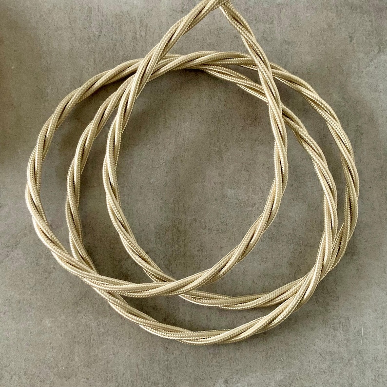 May include: A coiled, gold-colored electrical cord with a twisted design. The cord is made of a woven material and is arranged in a circular pattern against a gray background. The cord appears to be new and unused.