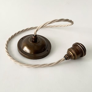 Puede incluir: Una lámpara colgante de latón de estilo vintage con un cable trenzado de color crema. La lámpara incluye una roseta de techo redonda y un portalámparas. El latón tiene un acabado cálido y envejecido, y el cable mide unos 76 cm.
