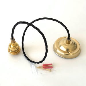 Puede incluir: Un colgante de lat&oacute;n con un cable trenzado negro. El conjunto incluye un portal&aacute;mparas, una roseta de techo y un peque&ntilde;o paquete con tornillos y tacos rojos. Sobre un fondo blanco.