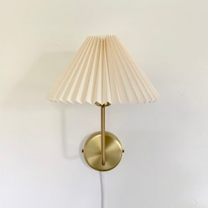 May include: A gold wall sconce with a white pleated fabric lampshade. The sconce has a round base and a single arm that extends upwards to support the lampshade.