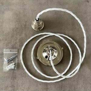 Op de afbeelding: Een plafondlamp kit met een zilverkleurige kap en een bijpassende decoratieve dop. De kit bevat een witte, gedraaide, met stof bedekte kabel en een klein zakje met schroeven en pluggen. De componenten zijn gerangschikt op een grijze ondergrond.