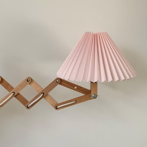 Puede incluir: Una l&aacute;mpara de pared de madera con un brazo flexible y una pantalla de l&aacute;mpara rosa plisada.