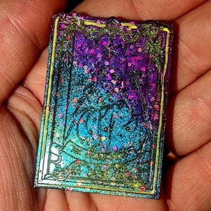 May include: A small, iridescent, rectangular card with a glittery, holographic finish. The card features a design of a figure with a crown, surrounded by a decorative border. The card is a vibrant mix of purple, blue, and green.