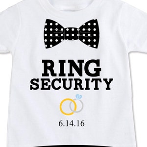 K&ouml;nnte beinhalten: Wei&szlig;es T-Shirt mit einer schwarz-wei&szlig;en Polka-Dot-Fliege, dem Text "RING SECURITY" und zwei goldenen Ringen mit einem Diamantring. Das Datum "6.14.16" ist unter den Ringen gedruckt.