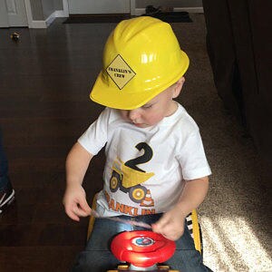 May include: A yellow construction worker hat with a black and yellow sign that says "Franklin's Crew". A young child is wearing the hat and a white t-shirt with a yellow dump truck and the number "2" on it.