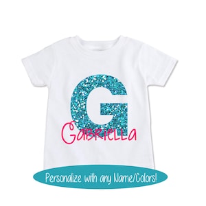 May include: White t-shirt featuring a large blue glitter letter G and the name "Gabriella" written in pink underneath.  Personalise with any name and colours.