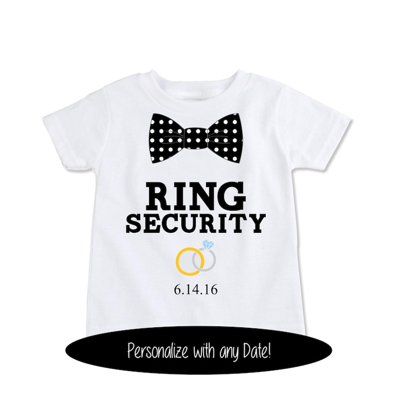 K&ouml;nnte beinhalten: Wei&szlig;es T-Shirt mit einer schwarz-wei&szlig;en Polka-Dot-Fliege, dem Text "RING SECURITY" und zwei goldenen Ringen mit einem Diamantring. Das Datum "6.14.16" ist unter den Ringen gedruckt.