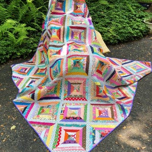 May include: A colorful handmade quilt with a patchwork design. The quilt features a variety of vibrant fabrics in squares and rectangles, with a purple border. The quilt is draped over a chair outdoors, showcasing its intricate patterns and textures.
