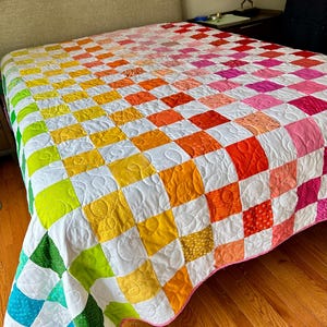 以下が含まれることがあります： 青からピンクへのグラデーションカラーの市松模様のカラフルなキルト。キルトは、ニュートラルなヘッドボードと木製の床のあるベッドの上にあります。