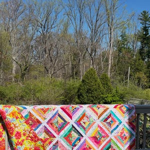 May include: A colorful handmade quilt with a geometric pattern of squares and triangles in various bright colors. The quilt is draped over a wooden railing outdoors, with a vibrant floral pattern on the reverse side.