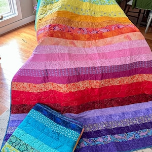 May include: A vibrant, rainbow-colored quilt with horizontal stripes of various colors, including red, orange, yellow, green, and blue. A smaller, folded quilt in shades of blue and green is also present. The quilts are likely handmade.