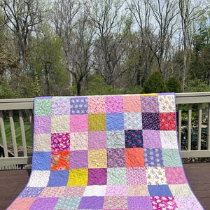 May include: A colourful patchwork quilt with a variety of squares. The quilt features floral, paisley, and solid fabric squares in shades of pink, purple, orange, and blue. The quilt has a purple border.