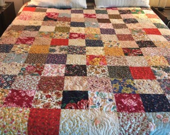 May include: A patchwork quilt featuring a variety of floral and patterned fabrics in red, yellow, blue, and other colours. The quilt is on a bed with two white pillows. The quilt is approximately 203 cm wide and 229 cm long.