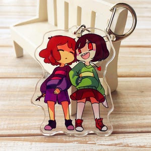Może przedstawiać: Przezroczysty brelok z akrylu przedstawiający dwie postacie z kreskówek z rudymi i brązowymi włosami, ubrane w zielone, fioletowe i czerwone ubrania. Jedna postać ma nóż. Brelok ma srebrne zapięcie.