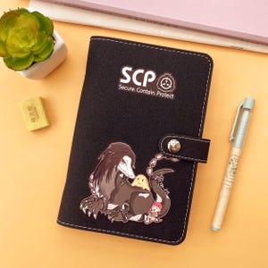Op de afbeelding: Zwart notitieboek met de tekst "SCP Secure. Contain. Protect" en een afbeelding van een wezen. Het notitieboek heeft een drukknoopsluiting en witte stiksels. Een pen en een kleine vetplant zijn ook zichtbaar.