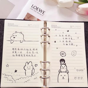 Op de afbeelding: Open notitieboek met gelinieerde pagina's, met zwarte inktkrabbels van een beer, een konijn en opgestapelde dieren. Handgeschreven Chinese karakters en Engelse tekst zijn ook aanwezig.