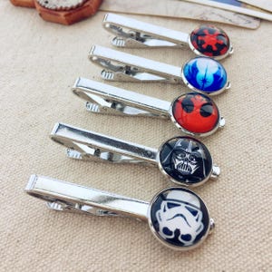 May include: Five silver-toned metal tie clips arranged in a row. Each clip features a circular glass cabochon top displaying different graphic designs: a red and black imperial symbol, a blue light symbol, a red mask, a black Darth Vader helmet, and a white Stormtrooper helmet.