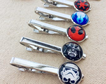 May include: Five silver-toned metal tie clips arranged in a row. Each clip features a circular glass cabochon top displaying different graphic designs: a red and black imperial symbol, a blue light symbol, a red mask, a black Darth Vader helmet, and a white Stormtrooper helmet.