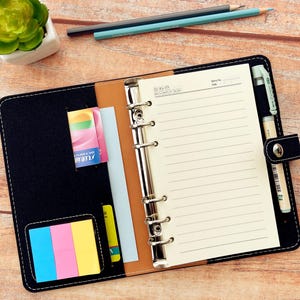 Op de afbeelding: Zwart leren planner met gelijnd papier, een pen en een pennenhouder. De planner heeft een drukknoopsluiting en een vak voor kaarten en plaknotities. Een kleine vetplant en potloden staan op de achtergrond.